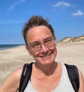 En leende person med kort brunt hår och glasögon i bakgrunden blå himmel och vit strand.
