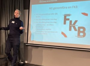 En person framme vid en scen pekar mot en ljus skärm med texten FKB