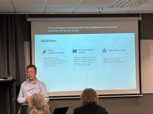 En person pratar inför en publik och vid sidan ser man en ljus skärm med text.