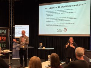 En person pratar inför en publik och har en presentation om Funktionsrättskonventionen i bakgrunden