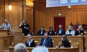 En person står i talarstol oh åtta personer sitter i bänkar bredvid varandra