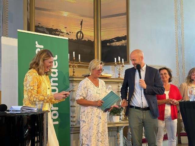 Fem personer står på en scen, en talar i mikrofon och ett par tittar leende ned på papper i handen.